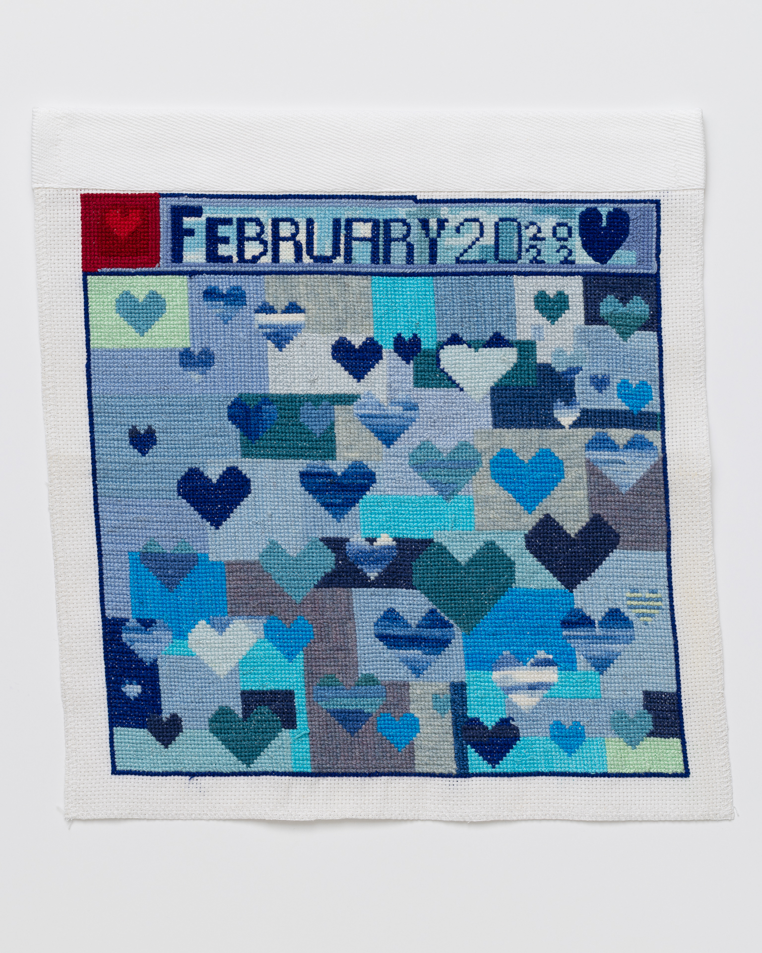 Cross stitch embroidery with various colored blue hearts on a background of vlue rectangles. "February 20 2022" is written across the top. There is a dark red square with a lighter red heart in the top left corner.