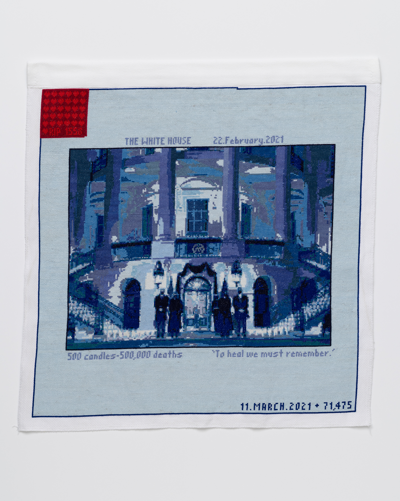 Cross stitch embroidery of picture of President Biden in front of the White House, standing with other figures, stitched in blue threads. There is a solid light blue border around the image, with "THE WHITE HOUSE 22.February.2021" written above, and "500 candles- 500,000 deaths To heal we must remember." written below. There is a red square in the upper left corner.