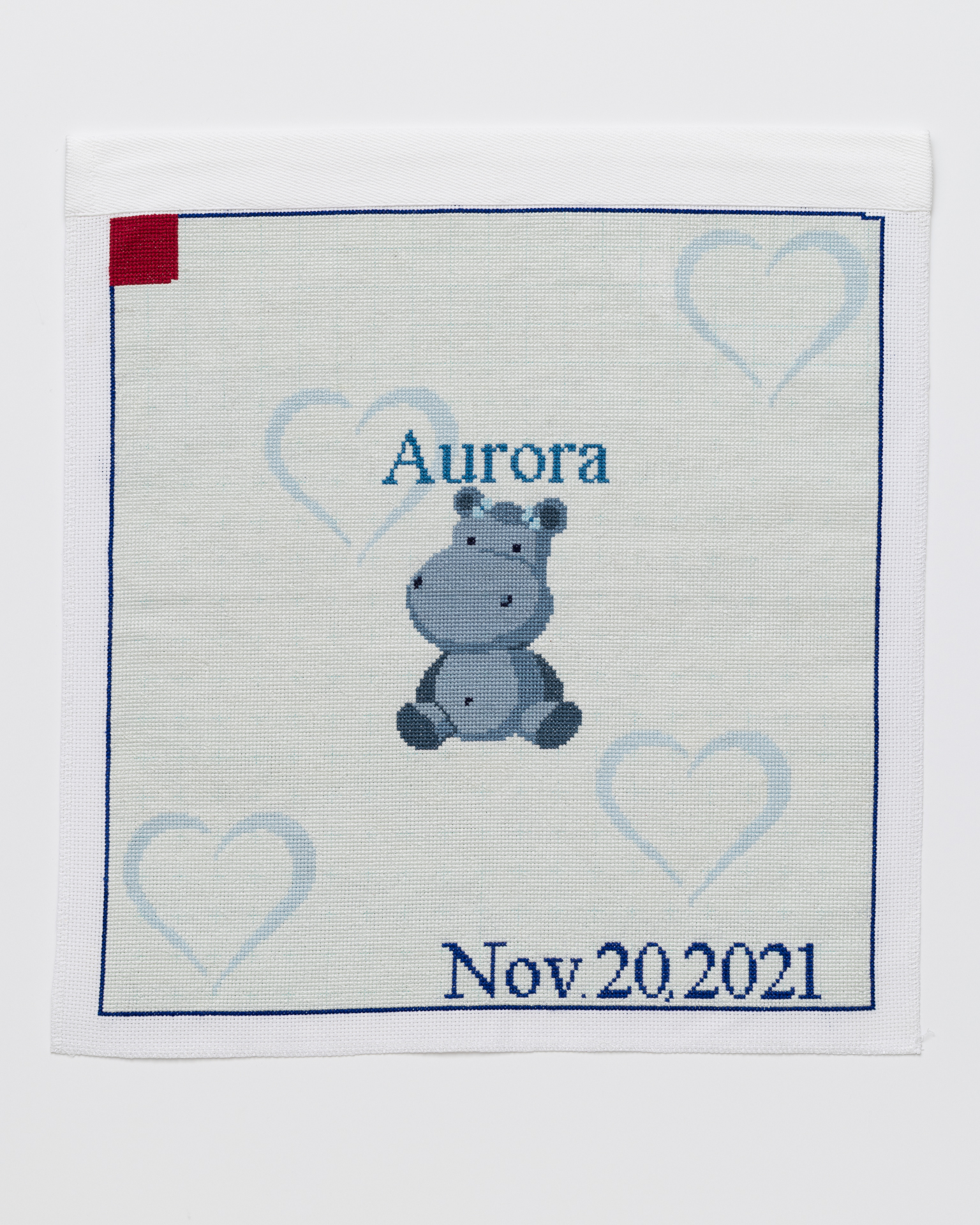 Nov 20, 2021 Cross stitch embroidery of a cartoon hippopotamus with the name "Aurora" stitched above it. The background is very light blue with light blue hearts. "Nov 20, 2021" is stitched on the bottom right corner, and a red square is in the top left.