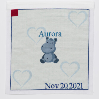 Cross stitch embroidery of a cartoon hippopotamus with the name "Aurora" stitched above it. The background is very light blue with light blue hearts. "Nov 20, 2021" is stitched on the bottom right corner, and a red square is in the top left.