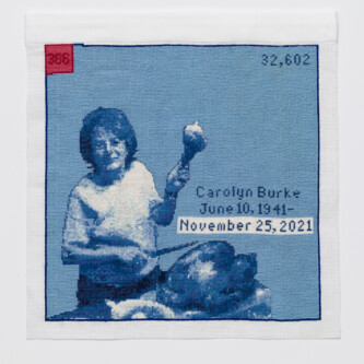 Cross stitch embroidery of a woman with short hair in a plain t-shirt holding up a turkey leg in her left hand, with a knife in her right hand. A cooked turkey sits on a table in front of her. Stitched in blue threads, with a red square in the top left corner. Across the middle right of the image is written "Carol Burke June 10, 1941 - November 25, 2021"