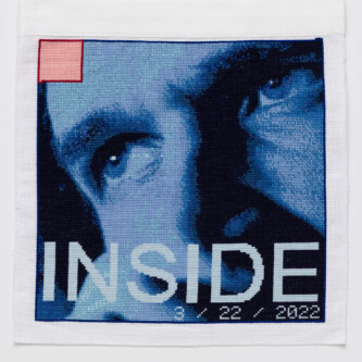 Photo by Wes Magyar (es translation) Cross stitch embroidery of the top half of a man's face in various colors of blue, with the eyes looking left. There is a small pink square outlined in red thread in the top left corner. Across the bottom of the image is the word "INSIDE" in light blue thread.