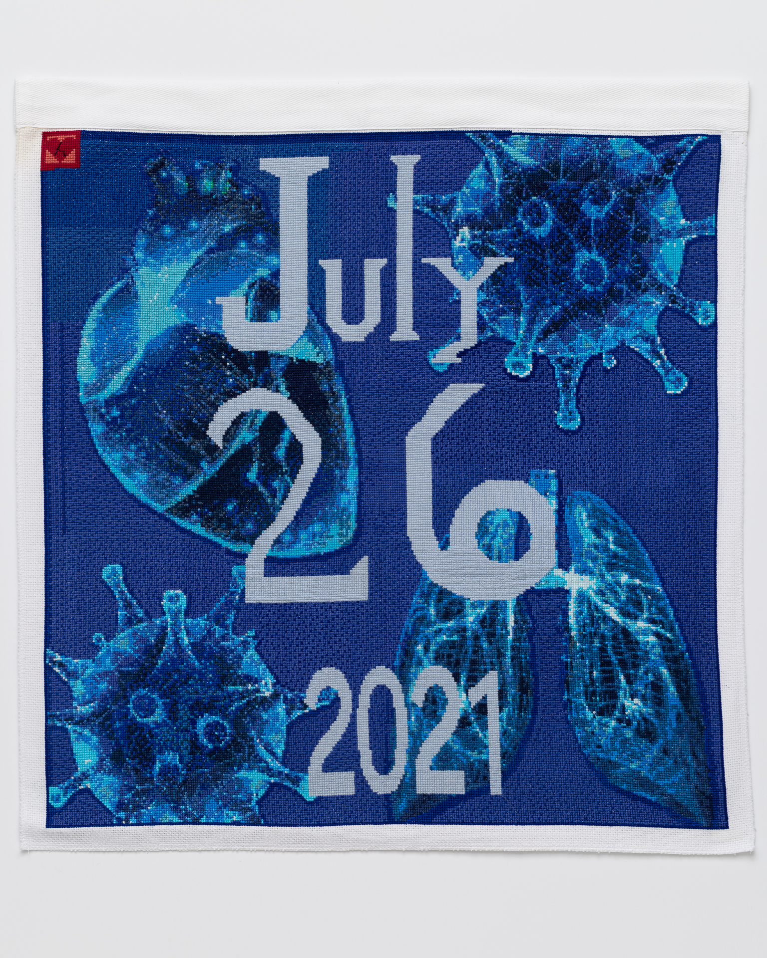 July 26, 2021 Cross stitch embroidery with "July 26 2021" down the middle. A human heart, two COVID virus images, and a set of lungs are embroidered in blue threads in behind the lettering. The background is royal blue. A red square is stitched in the top left corner with a small heart shape inside.